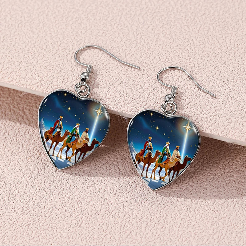 Heart-shaped earrings with a design of three wise men on camels and a star in the sky, displayed on a light pink background.